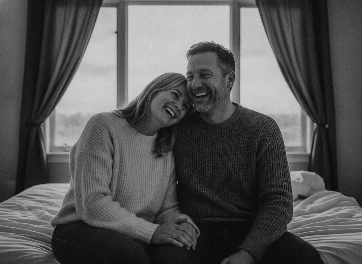 A couple in their forties, fully dressed in soft sweaters, sitting close on the edge of an unmade bed by a large window 